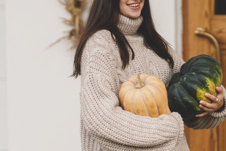 Happy woman in knitted sweater holding big autumn pumpkins for house entrance decor. Stylish woman...