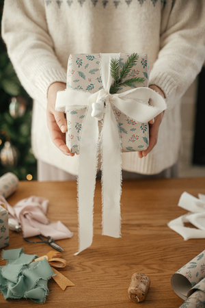 Woman holding stylish decorated christmas gift close up on background of table with festive...