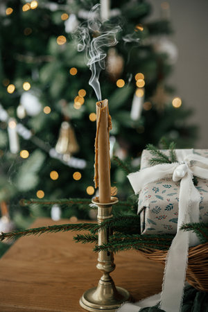 Stylish candle, wrapped christmas gift, rustic basket with fir branches against festive decorated tree in scandinavian room. Moody atmospheric image. Merry Christmas and Happy holidays!の写真素材