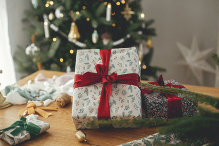 Stylish christmas gift with red ribbon close up on rustic table with festive decorations on background of modern decorated vintage tree in scandinavian room. Merry Christmas and Happy holidays!の写真素材