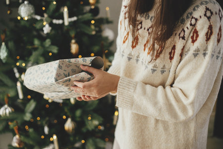 Woman in cozy sweater wrapping stylish christmas gift in paper on background of stylish christmas tree in decorative scandinavian room. Merry Christmas! Hands packing presentの写真素材