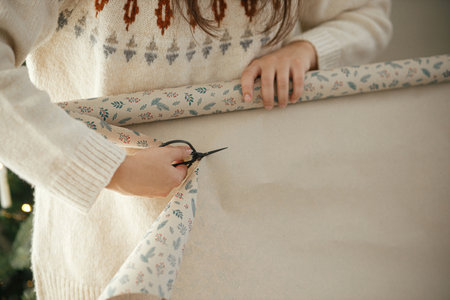 Woman cutting with scissors stylish festive paper for wrapping christmas gift close up in modern decorated scandinavian room. Winter holidays preparationの写真素材