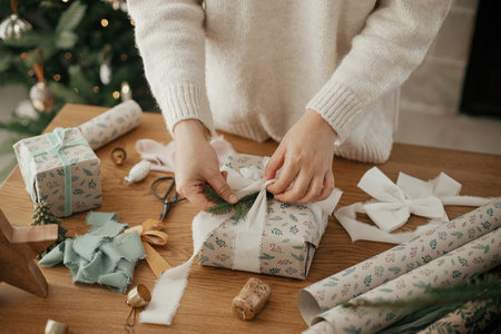 Woman wrapping stylish christmas gift with ribbon and fir branch on wooden table with festive decorations in modern decorated scandinavian room. Merry Christmas! Hands packing present close upの写真素材