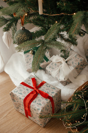Stylish wrapped christmas gifts and rustic basket with fir branches under festive decorated christmas tree in scandinavian room. Merry Christmas and Happy holidays!の写真素材