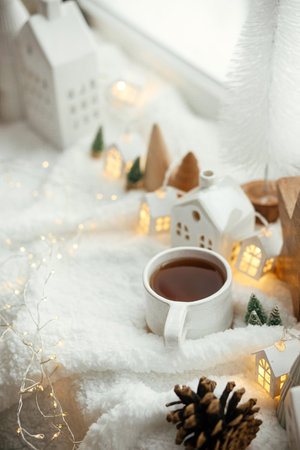 Winter hygge. Stylish cup of tea with modern cute christmas houses, pine cone, wooden star and tree, golden lights on soft warm blanket on windowsill. Christmas cozy still life. Merry Christmas!の写真素材