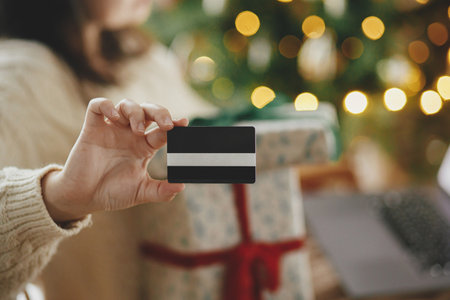 Woman in cozy sweater holding credit card close up and stylish christmas gifts in festive decorated christmas room with lights. Christmas shopping online and black friday sales.の写真素材