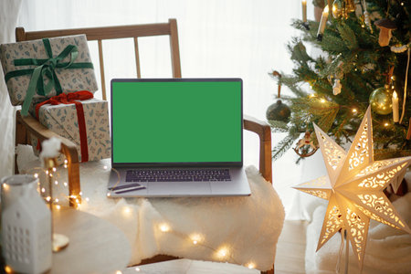 Laptop with empty screen and stylish christmas gifts on modern chair on background of festive decorated tree, golden lights and star in room. Christmas shopping online and sales. Laptop mock upの写真素材