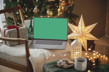 Christmas shopping online and sales. Laptop with empty screen and stylish christmas gifts on modern chair in festive decorated room with tree, golden lights and star. Laptop mock up, copy spaceの写真素材