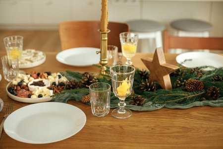 Christmas table setting. Stylish table runner with fir branches and pine cones, candles, cheese appetizers and starters, glasses and cutlery. Holiday dinner arrangement. Happy new year!の写真素材