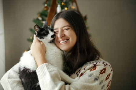 Happy woman in cozy sweater hugging cute cat on background of stylish decorated christmas tree in festive room. Owner with pet and winter holidays. Merry Christmas!の写真素材