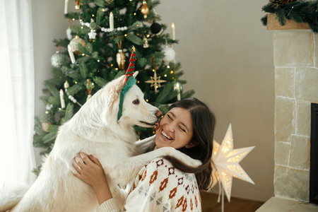 Happy woman in cozy sweater hugging cute white dog in festive accessory on background of stylish christmas tree. Winter holidays. Owner and pet playing in festive room. Merry Christmas!の写真素材