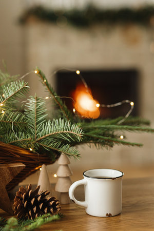 Stylish cup of warm tea, fir branches, wooden trees and star, pine cones on table against burning fireplace. Modern christmas rustic eco friendly decor and empty mug, scandinavian winter hyggeの写真素材