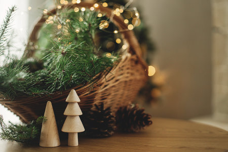 Stylish wicker basket with christmas fir branches, wooden trees, pine cones on table against burning fireplace. Modern rustic eco friendly decor for winter holidays in farmhouse living roomの写真素材