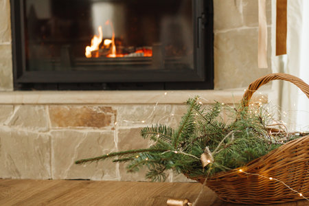 Rustic wicker basket with spruce branches, vintage bells and lights on background of burning fireplace. Festive christmas fireplace decor in modern farmhouse living roomの写真素材