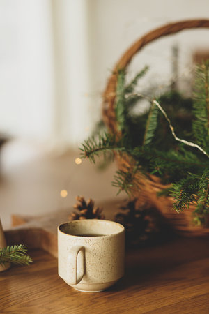 Winter holidays hygge. Stylish cup of warm tea, fir branches in basket, wooden trees and star, pine cones and lights on table. Modern christmas rustic eco friendly decor in scandinavian roomの写真素材