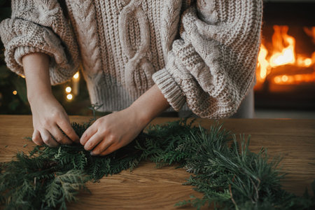 Hands in cozy sweater making Christmas rustic wreath with fir branches on wooden table against cozy fireplace, close up. Winter holiday preparations, atmospheric timeの写真素材