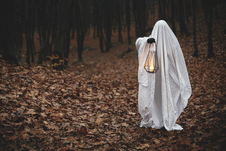 Spooky ghost holding glowing lantern in moody dark autumn forest. Person dressed in white sheet as ghost with light in evening fall woods. Happy Halloween! Trick or treatの写真素材