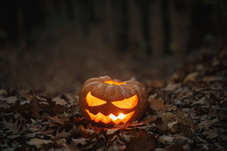 Spooky glowing Jack o lantern on autumn leaves in moody dark forest. Happy Halloween! Scary...