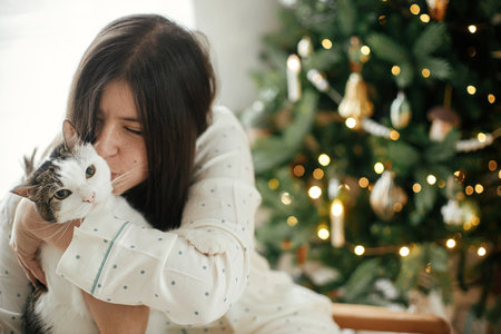 Happy woman in stylish pajamas hugging adorable cat on background of stylish christmas tree with lights in festive decorated living room. Merry Christmas! Winter holidays and petの写真素材