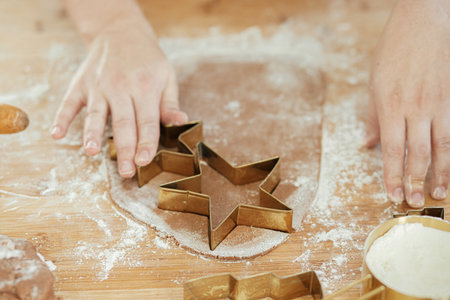 Hands cutting gingerbread dough with festive golden metal cutters on wooden board with flour,...