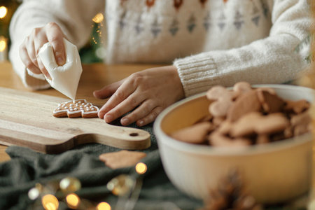 Hands decorating christmas gingerbread cookies with icing on rustic wooden table close up....