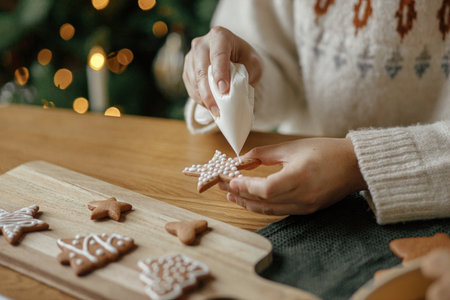 Hands decorating christmas gingerbread cookies with icing on rustic wooden table close up....