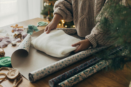 Wrapping christmas gifts. Hands in cozy sweater wrapping stylish sweater in festive wrapping paper with ribbons, vintage ornaments, bows on wooden table. Atmospheric winter holidaysの写真素材