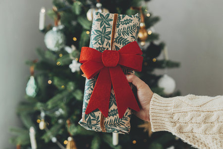 Merry Christmas! Woman in cozy sweater holding stylish christmas gift in festive wrapping paper with bow on background of christmas tree lights. Atmospheric winter holidays. Happy holidays!の写真素材