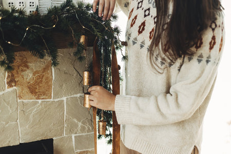 Decorating christmas fireplace with velvet ribbon and vintage bells on fir branches. Woman in cozy sweater hanging stylish decoration on mantel in modern farmhouse. Atmospheric winter holidayの写真素材