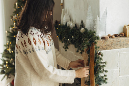 Decorating christmas fireplace with velvet ribbon and vintage bells on fir branches. Woman in cozy sweater hanging stylish decoration on mantel in modern farmhouse. Atmospheric winter holidayの写真素材