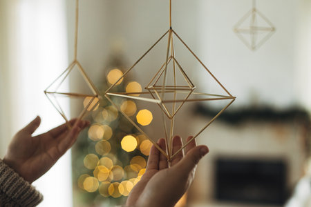 Hands in cozy sweater hanging rustic nordic straw decoration on background of festive golden lights...