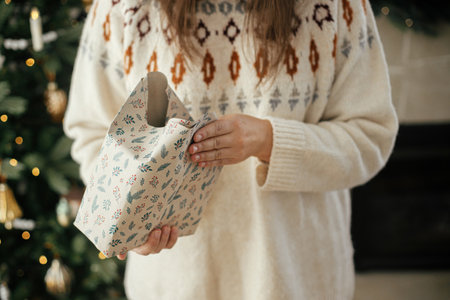 Woman in cozy sweater wrapping stylish christmas gift in paper on background of stylish christmas tree in decorative scandinavian room. Merry Christmas! Hands packing presentの写真素材