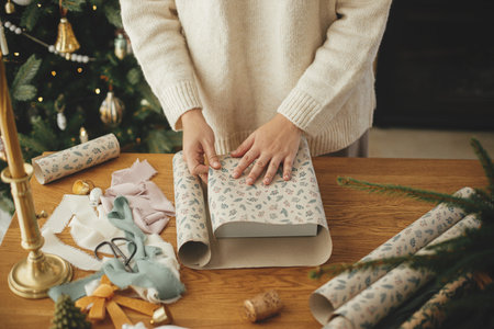 Woman in stylish sweater wrapping christmas gift in paper on wooden table with festive decorations in decorated scandinavian room. Merry Christmas! Hands packing present close upの写真素材