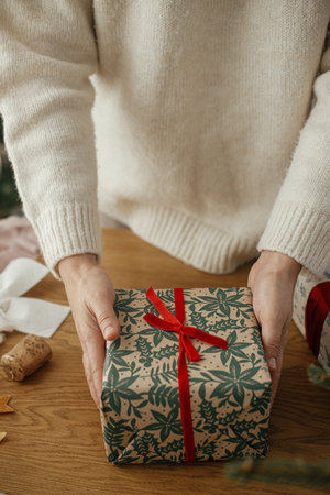 Woman holding stylish christmas gifts with red ribbon at wooden table with festive decorations in scandinavian room. Merry Christmas and happy holidays!の写真素材
