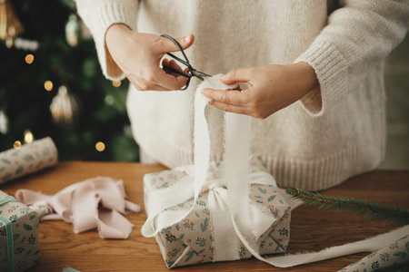 Woman in sweater wrapping christmas gift with stylish ribbon on wooden table with festive decorations in decorated scandinavian room. Merry Christmas! Hands packing present close upの写真素材