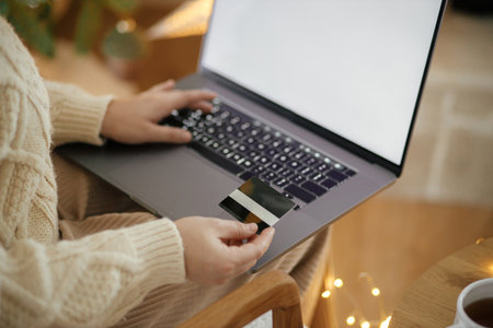 Christmas shopping online and black friday sales. Woman hands holding credit card on background of laptop with blank screen in festive decorated christmas room with lights. Space for textの写真素材