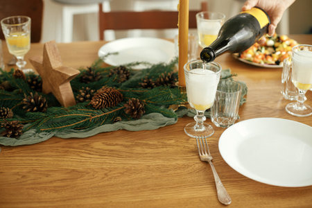 Christmas festive dinner. Hands pouring champagne into glasses on wooden table with fir branches and...