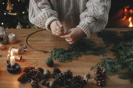 Hands in cozy sweater making Christmas rustic wreath with fir branches, pine cones, candle, twine, bells on wooden table, close up. Top view. Winter holiday preparations, atmospheric timeの写真素材