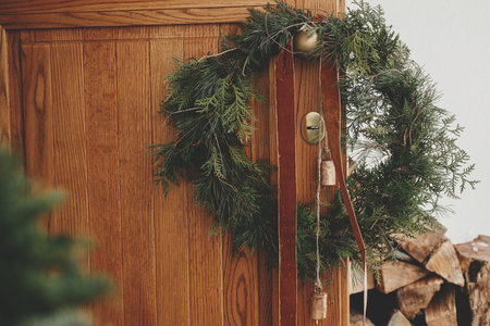 Stylish christmas rustic wreath with vintage bells and ribbon hanging on brass handle on wooden front door close up. Modern eco farmhouse exterior decor for winter rural holidaysの写真素材