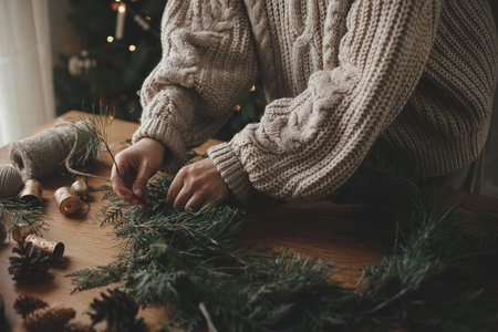 Making Christmas rustic wreath. Hands holding cedar branches, making wreath on wooden table with pine cones, candle, twine, bells in atmospheric festive room. Winter holiday preparationsの写真素材