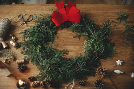 Stylish christmas rustic wreath with red bow, cedar branches, vintage bells, pine cones, twine, scissors on wooden table. Merry Christmas. Winter holiday preparations in atmospheric roomの写真素材