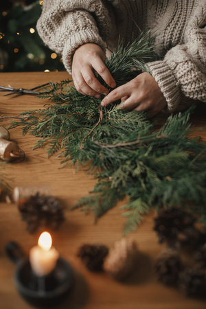 Making Christmas rustic wreath. Hands holding cedar branches, making wreath on wooden table with pine cones, candle, twine, bells in atmospheric festive room. Winter holiday preparationsの写真素材