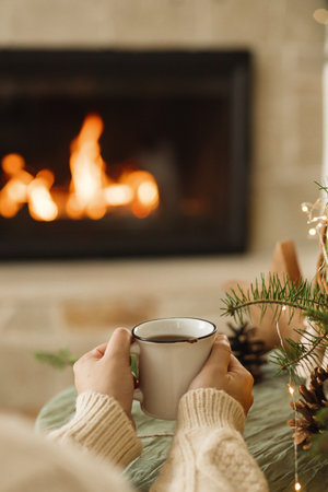 Hands in cozy sweater holding warm cup of warm tea on table with fir branches, wooden trees and and...