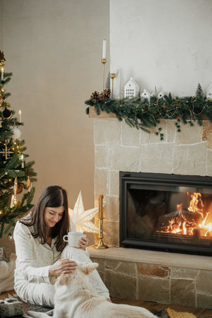 Beautiful woman in stylish pajamas with warm tea relaxing with cute dog at cozy fireplace, enjoying christmas morning in festive decorated living room with gifts. Merry Christmas! Winter holidaysの写真素材