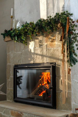 Stylish decorated christmas fireplace in minimal scandinavian living room. Modern christmas trees, houses, pine cones and spruce branches on fireplace mantel with brass bells and ribbon.の写真素材