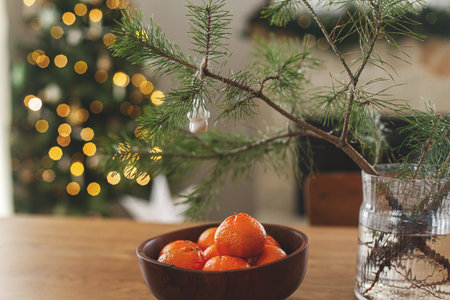 Fresh tangerines in wooden bowl and spruce branch on table against stylish decorated christmas tree with golden lights in scandinavian room. Atmospheric winter holidays at festive homeの写真素材