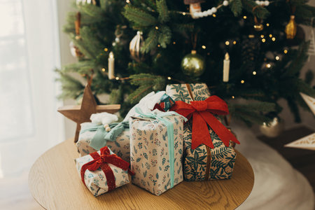 Stylish christmas gifts in festive wrapping paper with bows and vintage ornaments on wooden table on background of golden christmas tree lights.Happy holidays! Atmospheric christmas eveの写真素材