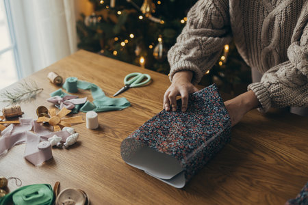 Wrapping christmas gifts. Hands in cozy sweater wrapping stylish present in festive wrapping paper with ribbons, vintage ornaments, bows on wooden table. Atmospheric winter holidaysの写真素材
