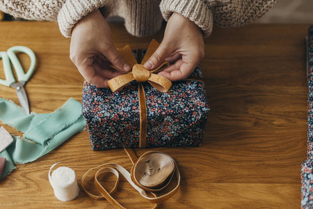 Merry Christmas! Woman in cozy sweater holding stylish christmas gift in festive wrapping paper with bow, ribbon, vintage ornaments on wooden table. Atmospheric winter holidaysの写真素材