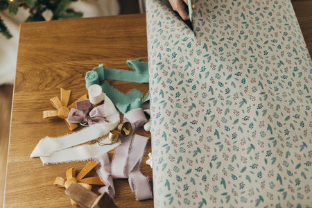 Wrapping christmas gifts. Hands in cozy sweater holding stylish festive wrapping paper with ribbons, vintage ornaments, bows on wooden table. Atmospheric winter holidaysの写真素材